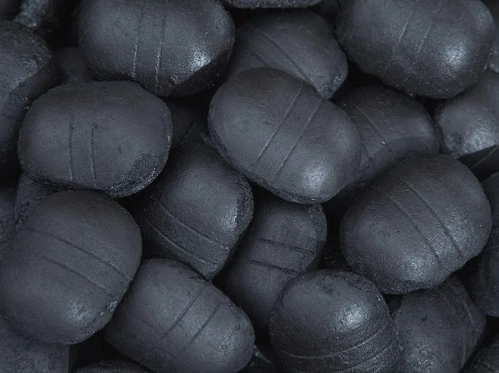 A dense heap of Super Blend smokeless ovoid briquettes, matte black with faint moulded seam lines and a fine grey dust bloom